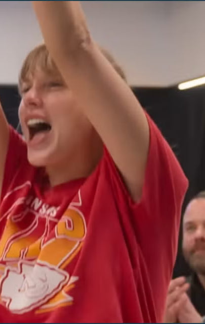 Taylor Swift in a JUNKFOOD red shirt with a graphic design, arms raised in an indoor setting