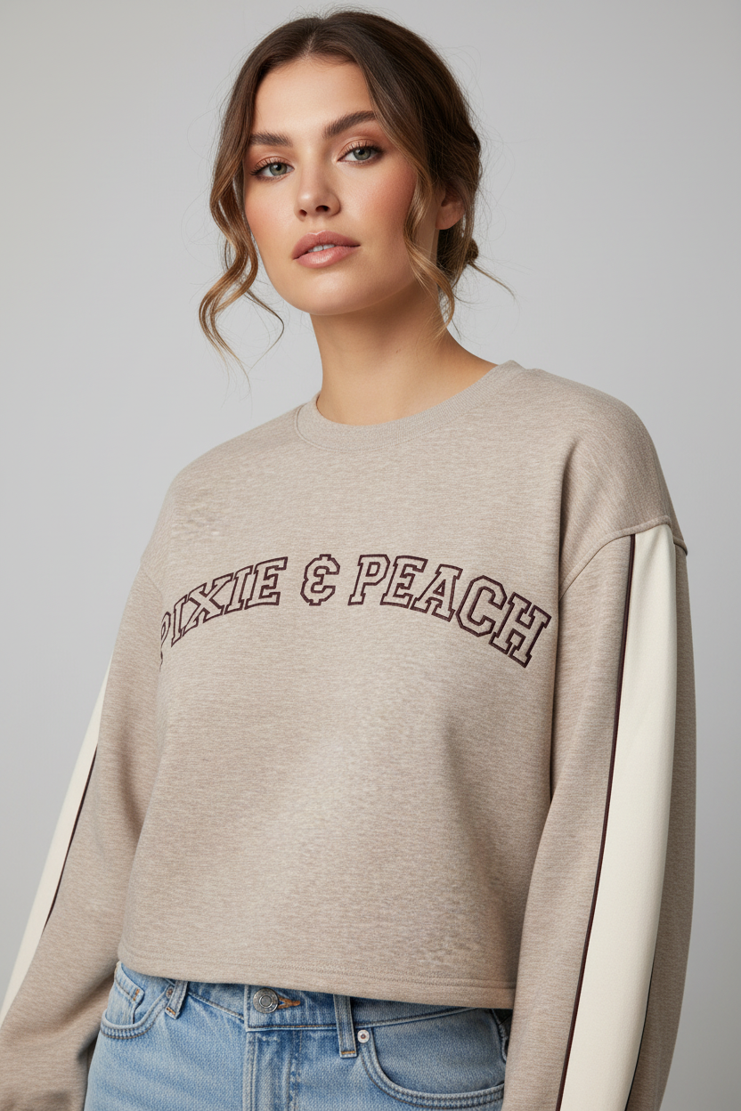 A cropped long-sleeve sweatshirt in a heathered beige color, featuring the words 'PIXIE & PEACH' arched across the chest in a dark brown outline. The sleeves have a contrasting solid light beige panel running down the side, and the cuffs and hem are ribbed.