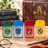 Four Harry Potter-themed hand sanitizer bottles in green, blue, red, and yellow, lined up on a wooden table with Hogwarts books, a leather trunk, glasses, and a wand in the background.