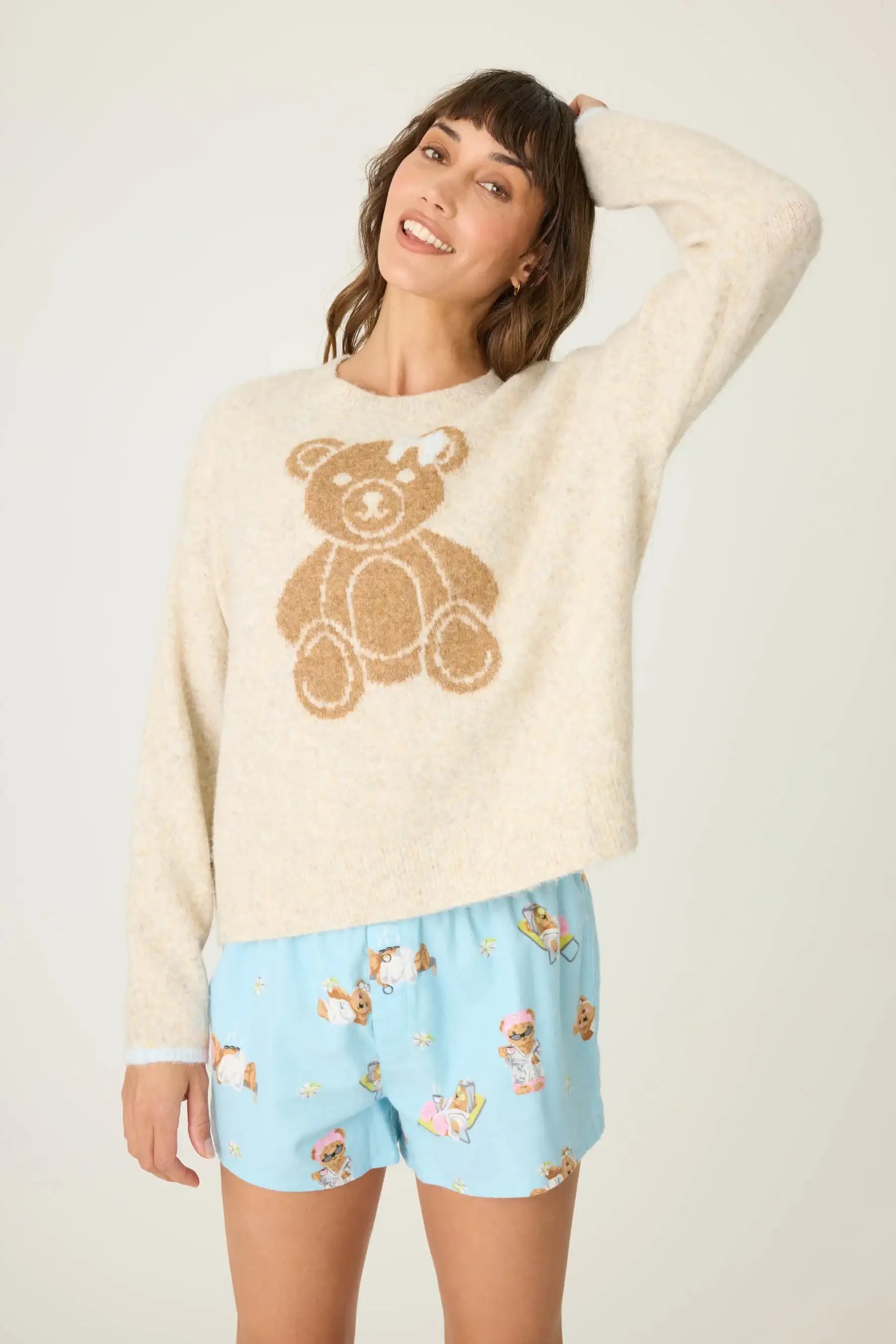 Woman wearing a cream sweater with a teddy bear design and light blue shorts with teddy bear pattern on a white background