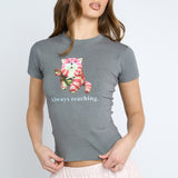 A woman wearing a heather gray short-sleeve t-shirt with a graphic of a pink and white cat holding a strawberry and the text 'Always reaching.' below it, paired with a light pink skirt, against a light gray studio background.