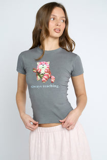 A woman wearing a heather gray short-sleeve t-shirt with a graphic of a pink and white cat holding a strawberry and the text 'Always reaching.' below it, paired with a light pink skirt, against a light gray studio background.